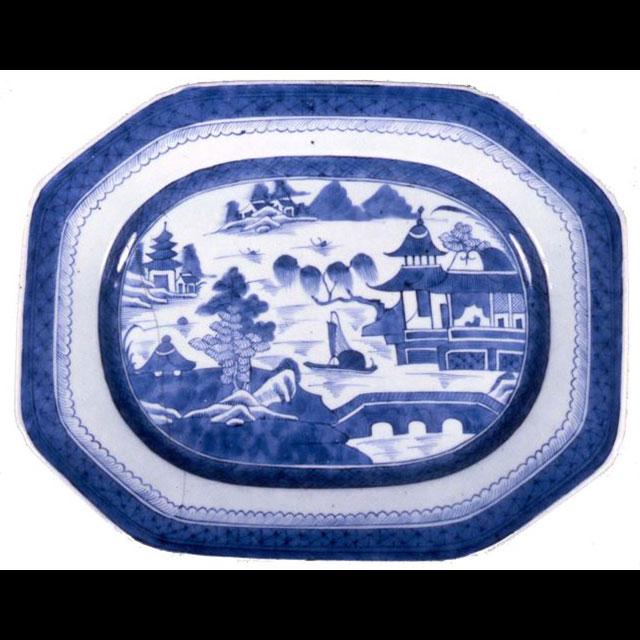 A blue-and-white ceramic platter made of Chinese porcelain. The border is dark blue with a crisscrossed motif, followed by a light blue, scalloped motif. The center is a scene of a river, with several pagodas on either side. There is a ship in the center and a bridge on the bottom right.