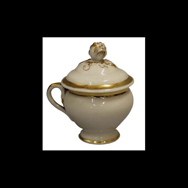A color photograph of a porcelain cup with a lid on top. The cup is white porcelain with gold detailing at the top of the lid, around the lip of the lid, around the base of the cup, and on the handle. There is a small filigree pattern in the center of the lid. 