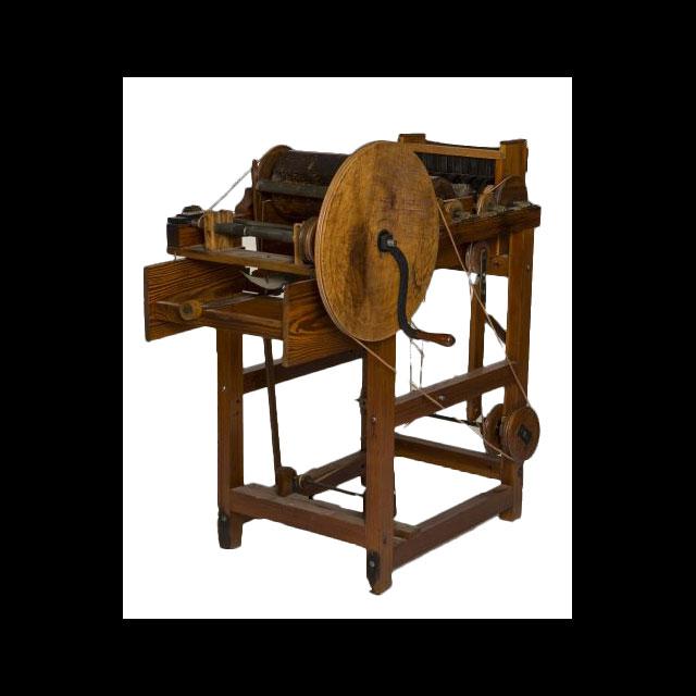 A color photograph of a wooden cotton gin. There is a large wheel on the right side to crank gears to clean cotton with drums connected by leather straps. The back of the gin curves into a chute. 