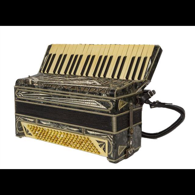A color photograph of Dominic Briagioli’s accordion. The keys and bottom design have yellowed. The case is black, with various patterns of faux jewels. A leather strap is attached to the back of the case.