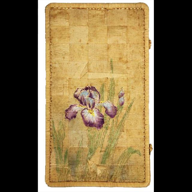 A color photograph of a straw handkerchief case. The case is yellow and the straw was woven in a patchwork pattern. In the center is a purple iris with blue centers in its petals. There are green leaves on either side. 