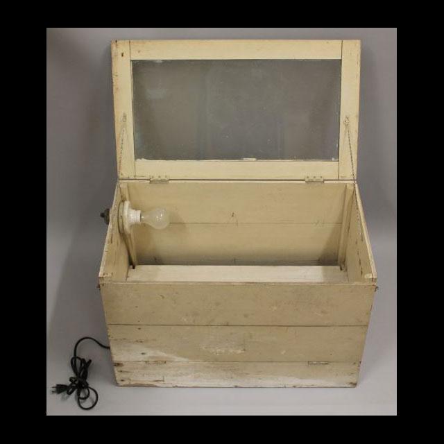A color image of a model baby incubator. The model is made of unfinished wood that has damage along the edges. There is a lightbulb in a white socket in the top left corner and a black cord attached to the left side. The lid is open, held by two chains. There is glass in the lid to observe the baby with the lid closed. 