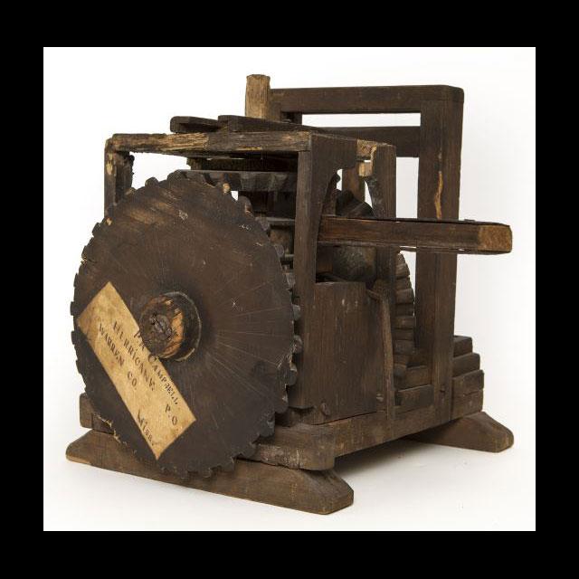 A color photograph of a patent model screw press. The model is made of wood and iron. The lever in the center moved the gears on the left, moving a vertical screw. There is a label attached to the gear.